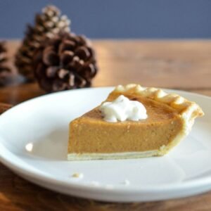 Close up of pumpkin pie on a white plate with out of focus pinecones in the back.