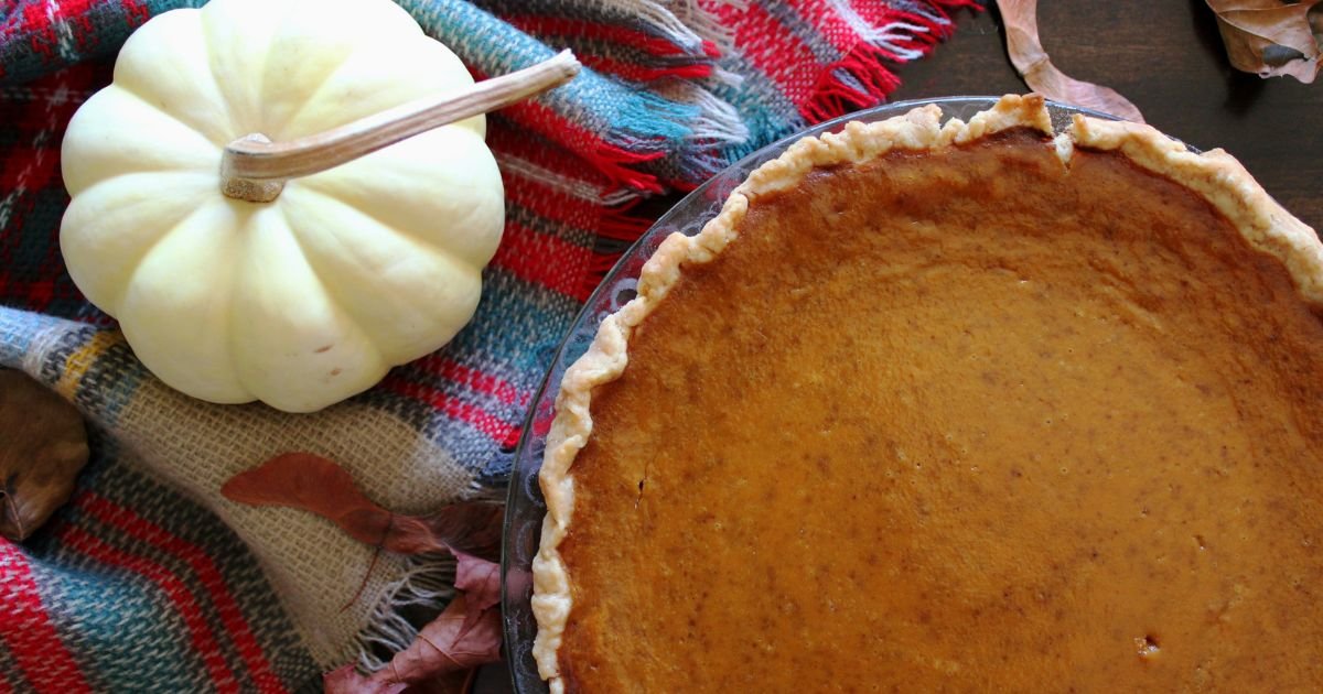 Overview of cooked pumpkin pie next to a small white pumpkin squash on top of woven fabric in a stripe pattern.
